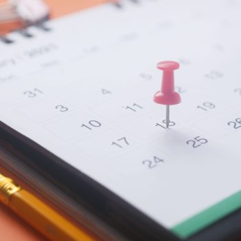 A close-up of a calendar page with a pink push pin, pencil, and binder clip on an orange background.